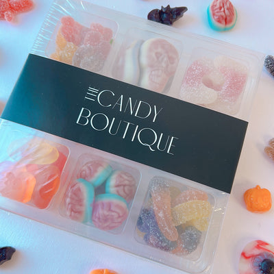 A clear plastic candy box with a black label reading 'The Candy Boutique'  surrounded by scattered Halloween-themed gummy candies, including orange pumpkins, pink skulls, and brain-shaped sweets and more.