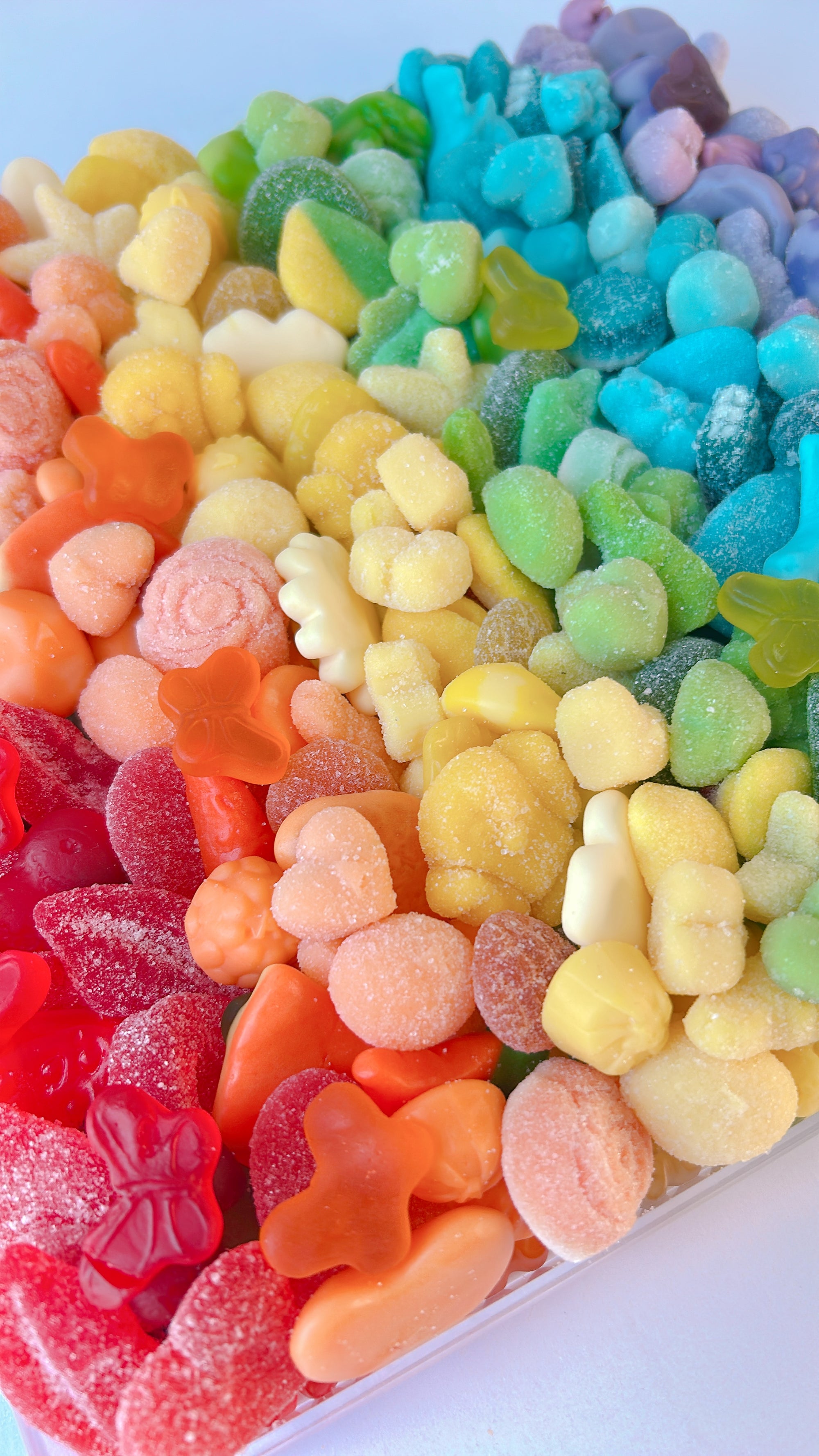 A close-up of a candy assortment in a clear container, arranged in diagonal rainbow rows of red, orange, yellow, green, blue, and purple gummy and sugar-coated candies.