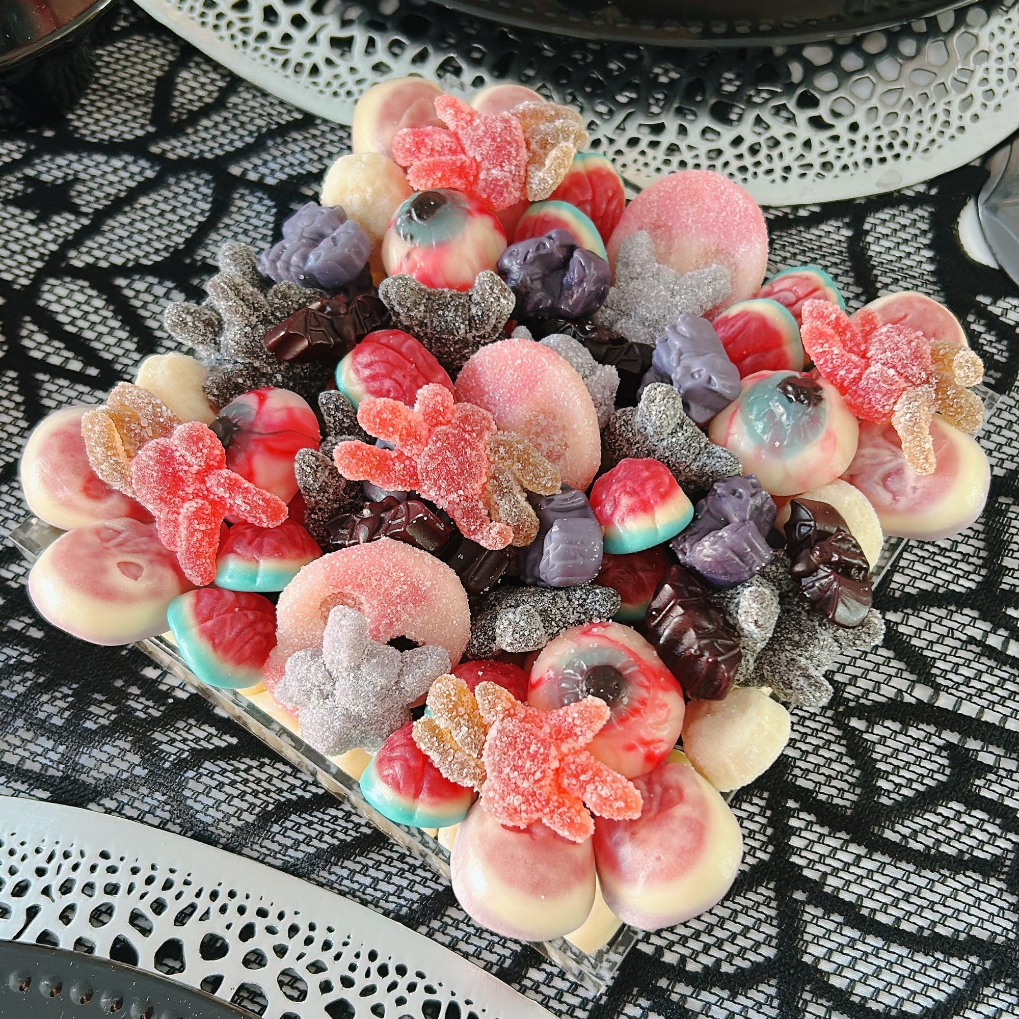 A close-up of a decorative arrangement of assorted colorful halloween-themed candies and gummy sweets placed on a black lace tablecloth, with plates partially visible around it.