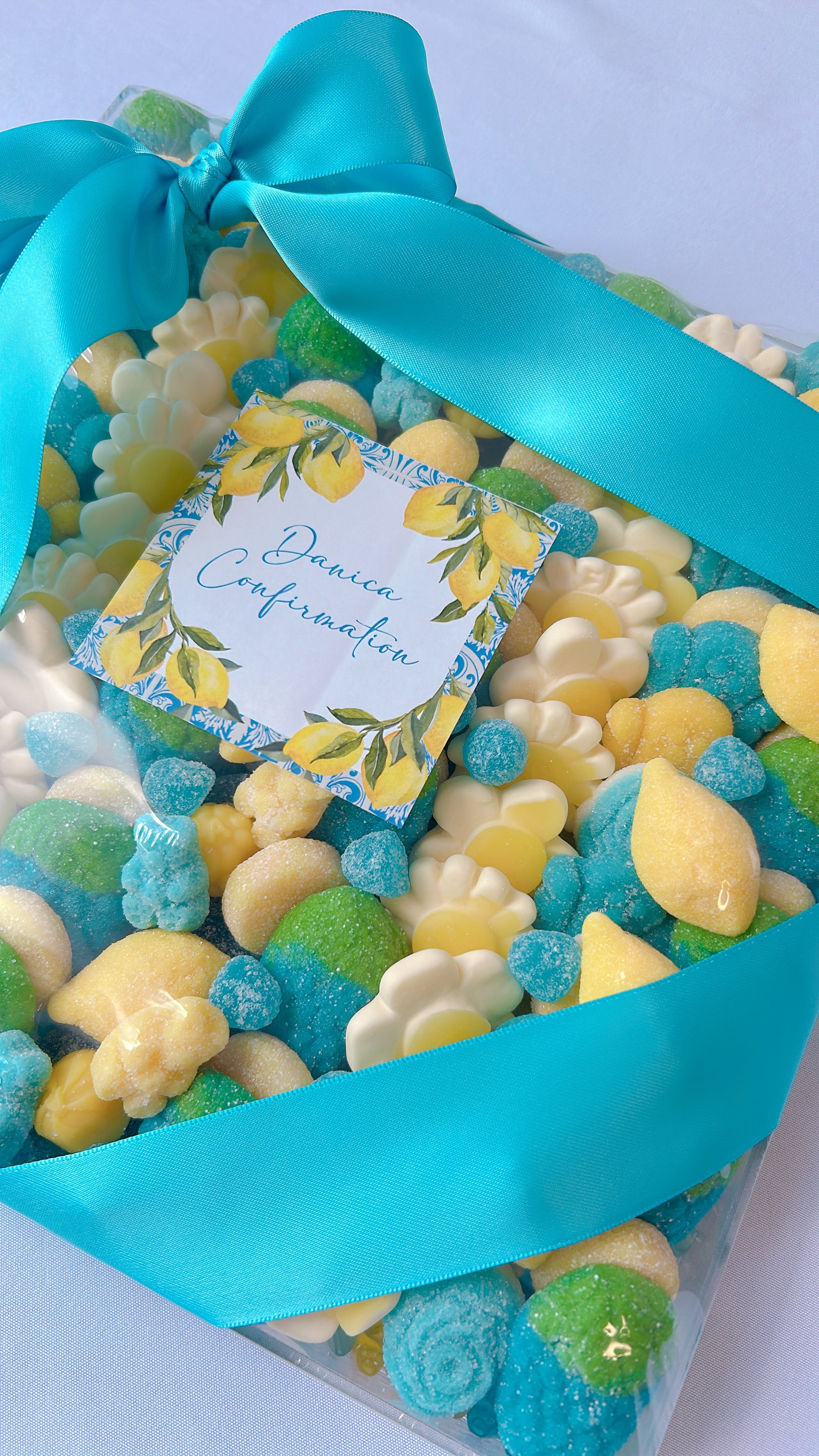A clear gift box filled with assorted colorful candies, tied with a bright turquoise ribbon and a small floral tag that reads 'Pinaud Confirmation.'.