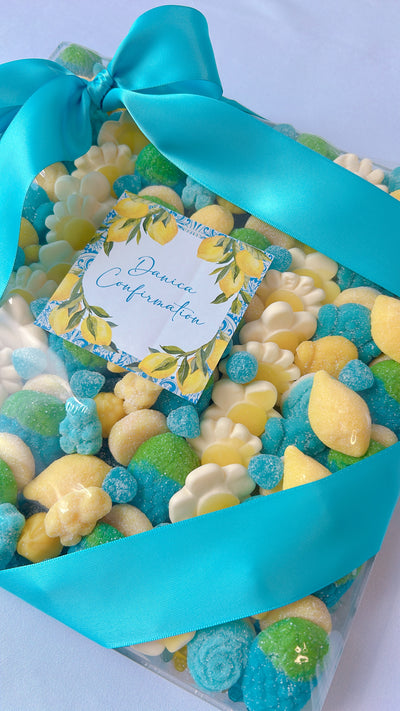 A clear gift box filled with assorted colorful candies, tied with a bright turquoise ribbon and a small floral tag that reads 'Pinaud Confirmation.'.
