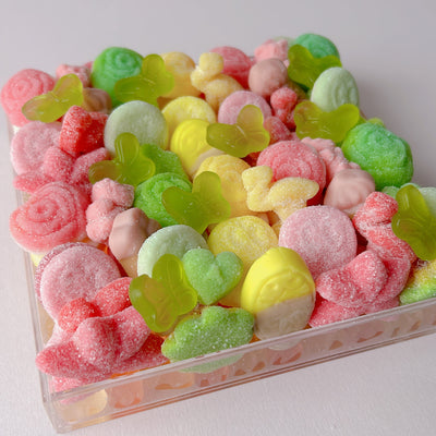 A clear acrylic container filled with assorted colorful gummy candies, including pink, green, yellow, and peach pieces, photographed on a plain light background.